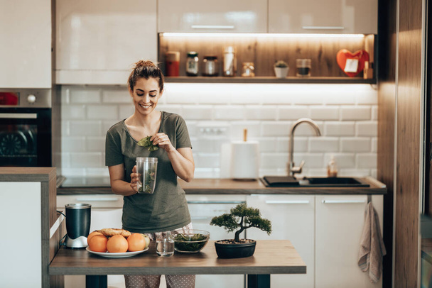 Young smiling woman putting a variety of  vegetables into a blender and making a healthy smoothie at home. - Photo, Image