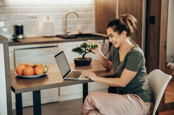 Tiro de uma mulher usando seu laptop e desfrutar de café da manhã em sua cozinha pela manhã em casa. - Foto, Imagem