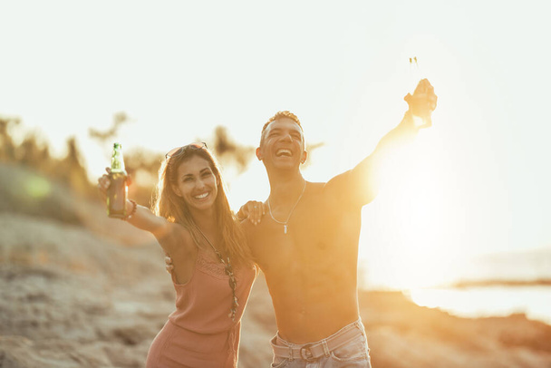 Young couple dancing and drinking beer on the beach by the sea. Enjoying in beautiful sunset and their love. - Zdjęcie, obraz