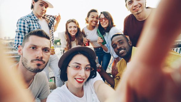 Mujer joven se está tomando selfie sosteniendo la cámara y mirando a la cámara mientras sus amigos están tintineando gafas, posando y mostrando gestos de la mano en la divertida fiesta al aire libre
. - Foto, imagen