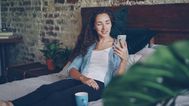 Good-looking woman is talking to friend online using modern smartphone making video call, she is happy and excited. Young lady is sitting on bed at home wearing casual clothes. - Fotografie, Obrázek