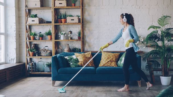 Cheerful young woman is dancing with mop during routine clean-up in nice loft style apartment, she is listening to music with wireless headphones and singing enjoying her occupation. - Foto, imagen