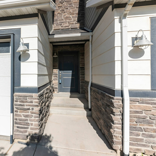 Square Dark gray front door in the middle of townhouses with stones and vinyl wood sidings. Entrance in the middle of garages with wall lamps and white garage doors with window panels. - Photo, Image