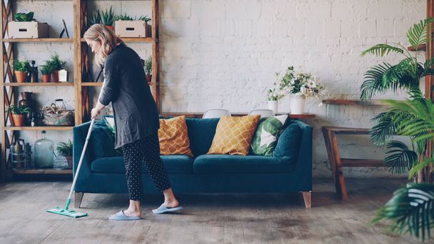 Attractive young woman is cleaning living room mopping the floor doing housework. Beautiful loft style apartment with modern furniture and green plants is visible. - Valokuva, kuva