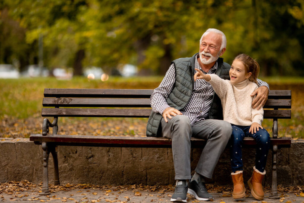 Красивый дедушка проводит время со своей внучкой на скамейке в парке осенью - Фото, изображение