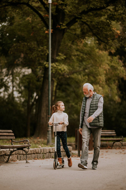 Senior man teaching his granddaughter how to ride kick scooter in the autumn park - Fotografie, Obrázek