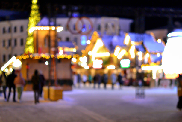 Blurred background. People walk in city square on winter night. Black silhouettes of people walking near houses decorated luminous illumination. White light bokeh blur spots from glowing house lights - Photo, Image