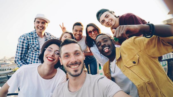 Point of view shot of bearded young man looking at camera taking selfie with friends sitting on roof at table and having fun. - Фото, изображение