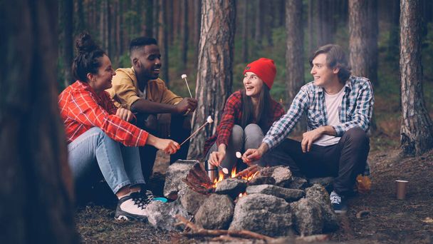 Hungry travelers are cooking marshmallow on fire and eating it from sticks during conversation around campfire, young people are talking and laughing enjoying sweet food. - Foto, imagen