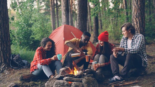 Handsome African American man is pouring hot drink from thermos bottle into glasses and cups sitting around fire at camp with happy friends multiethnic group. - 写真・画像