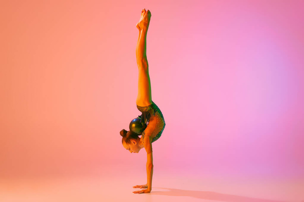 Studio shot of young charming girl, rhythmic gymnast training with sports equipment isolated over pink background in neon light filter. Dance, music, emotions, challenges, motivation and ad - Fotografie, Obrázek