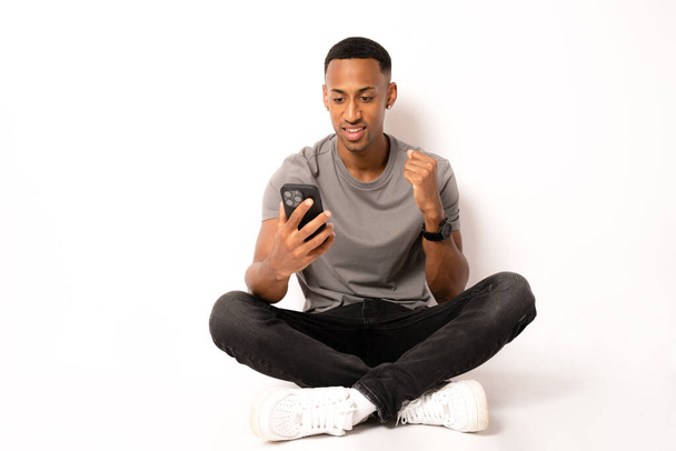 Молодой африканский американец сидит на полу и работает в смартфоне на белом фоне. - Фото, изображение