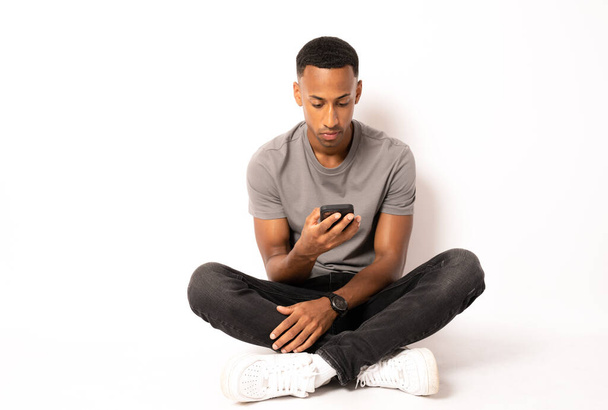 Młody Afroamerykanin siedzi na podłodze i pracuje w smartfonie na białym tle. - Zdjęcie, obraz