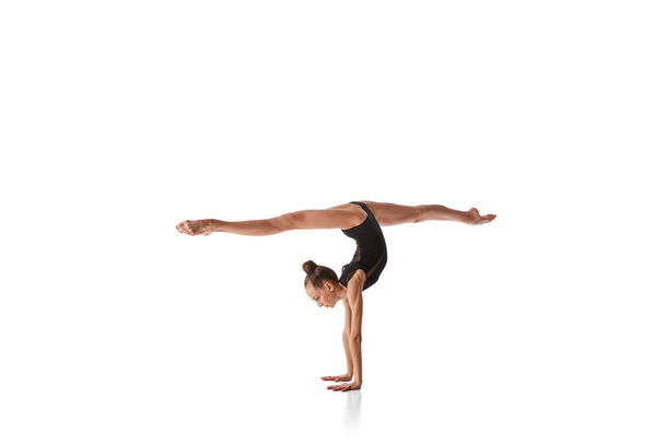 Twine in hand stand. Portrait of junior gymnast in black sport swimsuit doing gymnastics excercises isolated over white background. Sport, skills, achievements - Fotó, kép