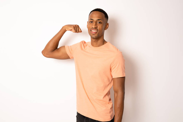 Молодий афроамериканець, одягнений в футболку, що стоїть над ізольованим білим фоном Сильний чоловік з м'язом руки, впевнений і гордий за владу. - Фото, зображення