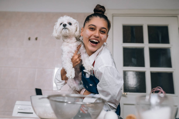 Sorrindo jovem mulher na cozinha segurando um cão maltês branco bonito - Foto, Imagem