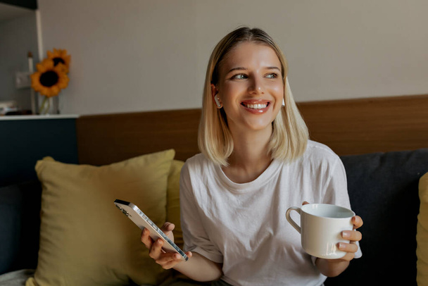 incredible cute lovely blond woman with short hairstyle wearing white t-shirt is sitting on sofa with smartphone and coffee and looking at window.  - Photo, Image