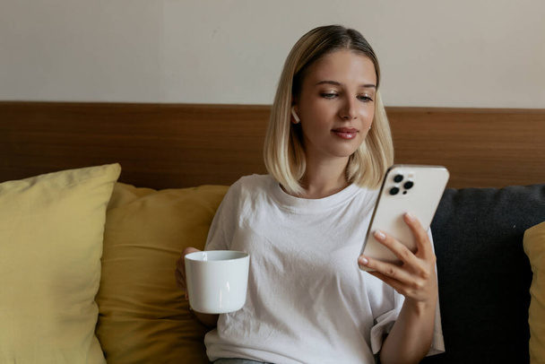 Indoor close-up portret van charmante mooie vrouw met blond haar dragen witte t-shirt zitten met koffie in hoofdtelefoon met smartphone - Foto, afbeelding