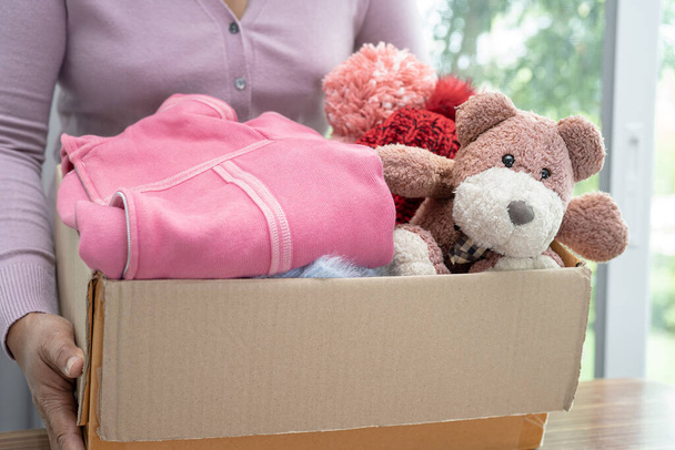 Κρατώντας κουτί δωρεά ενδυμάτων με μεταχειρισμένα ρούχα και κούκλα στο σπίτι για την υποστήριξη της βοήθειας για τους φτωχούς ανθρώπους στον κόσμο. - Φωτογραφία, εικόνα