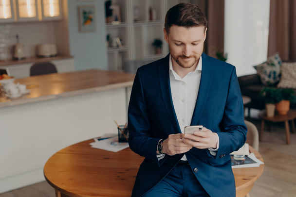 Confident bearded businessman in stylish suit leaning on table in living room and reading messages or last news in internet while holding smartphone. Business people lifestyle concept - 写真・画像