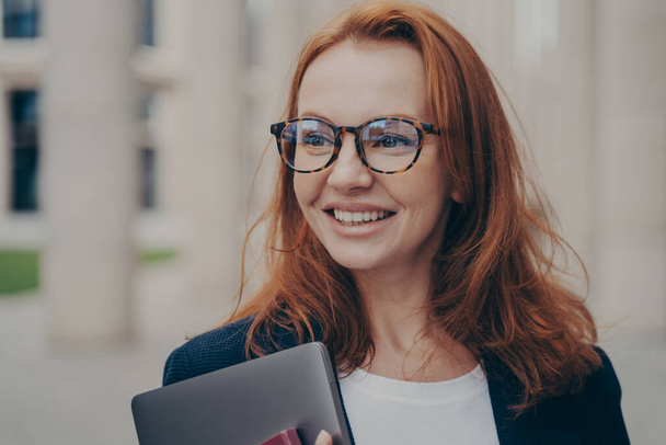 Portret szczęśliwej eleganckiej kobiety z szerokim uśmiechem patrząc na zewnątrz, czekając na partnera biznesowego na świeżym powietrzu, trzymając nowoczesny laptop i nosząc okulary, ruda kobieta biznesu wraca do domu po pracy - Zdjęcie, obraz