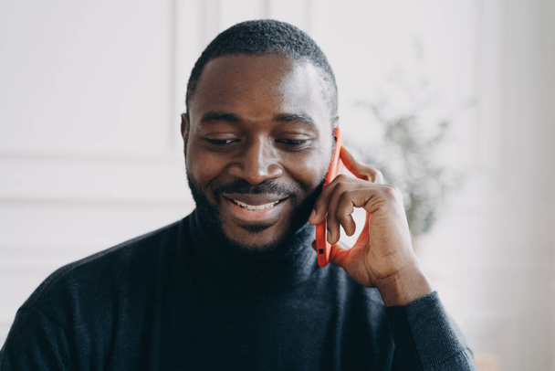Close up of young smiling african man having business conversation on mobile phone while working in office, cheerful dark-skinned guy holding smartphone talking with friend or consulting client - Photo, image