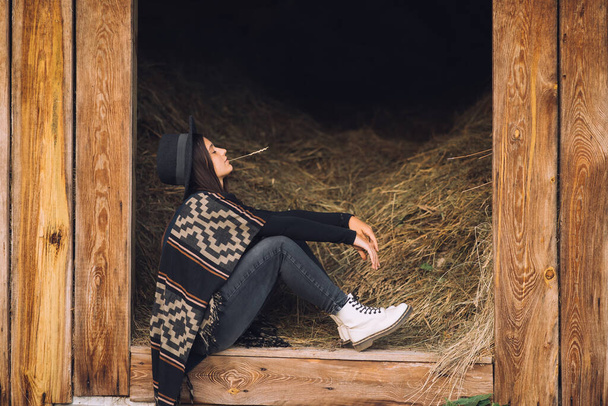 Young brunette woman poses while sitting at the entrance to the barn. Rural style. - 写真・画像