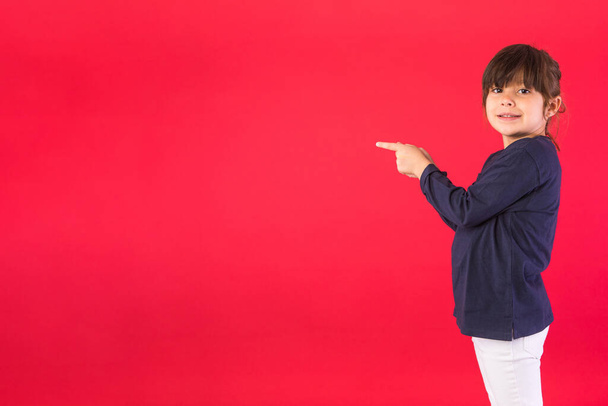 Smiling brunette haired girl, wearing blue sweatshirt and white pants, pointing fingers to the side with copy space, on red background. Concept of call center, call, speak and listen. - Photo, Image