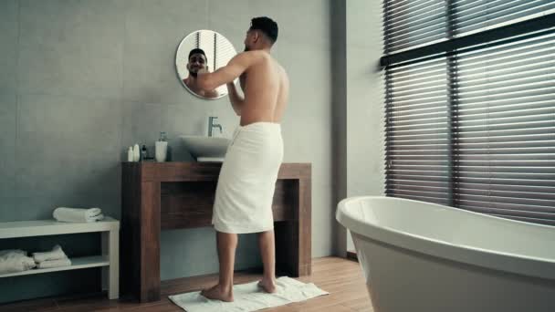 Full length view moving with hands dancing funny cheerful man with white bath towel wrapped around hips in bathroom looking in mirror pampering celebrate winning dance after shower having fun to music - Footage, Video