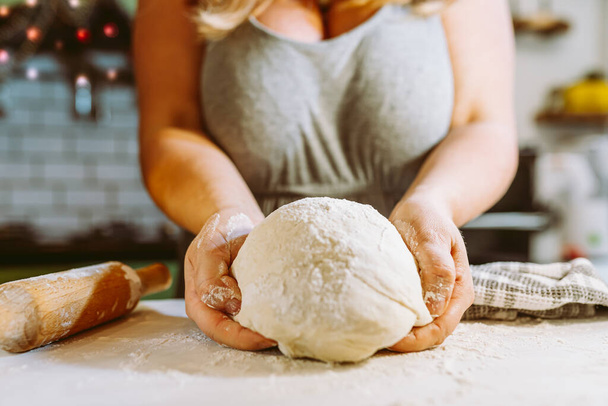 curvaceous woman, housewife, holds dough sprinkled with flour in hands, on kitchen table, against blurred background of kitchen decorated with Christmas tinsel. cooking dough for holiday baking, pizza, savory pie, at home, in home kitchen - Fotó, kép