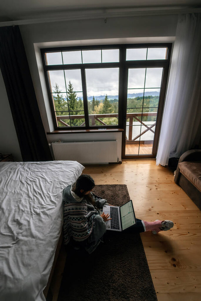 Drukke jonge vrouw met laptop op de vloer zitten door een tweepersoonsbed en netwerken of gewoon besteden vrije tijd op verschillende websites - Foto, afbeelding