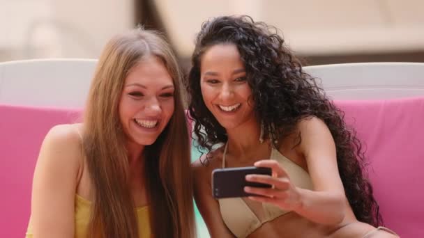 Όμορφη Ισπανίδα Καυκάσια κοπέλα φίλες γυναίκες με μπικίνι blogger ταξιδεύουν στο εξωτερικό κάνουν ηλιοθεραπεία στην ξαπλώστρα του θέρετρου spa χρησιμοποιούν βίντεο κλήση mobile εφαρμογή απολαμβάνουν ευχάριστη συνομιλία online μέσω τηλεφώνου - Πλάνα, βίντεο