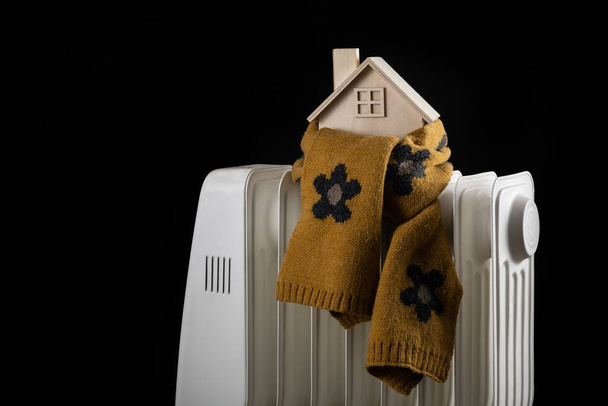 Envuelta en una cálida bufanda, una simbólica casa de madera se levanta sobre un radiador eléctrico. - Foto, Imagen