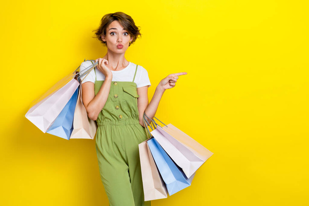 Photo of cute funky pouted lips lady wear khaki overalls trendy outfit finger directing new shopping mall sale isolated on yellow color background. - Фото, изображение