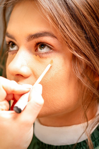 Bella giovane donna bruna che riceve il trucco con bastone correttore sul suo viso in un salone di bellezza - Foto, immagini