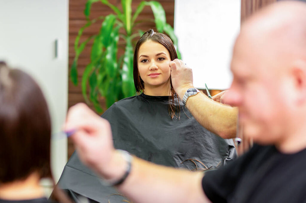若い白人女性の鏡の中に映る姿が、美容室で男性の美容師によるヘアカットを受けています。 - 写真・画像