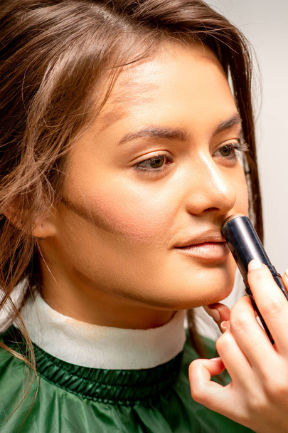Krásná mladá brunetka žena dostává make-up s tyčinkou maskér na její tváři v salonu krásy - Fotografie, Obrázek