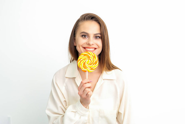 Beautiful young caucasian woman wearing a white shirt licking a lollipop on a white background - Foto, Imagen
