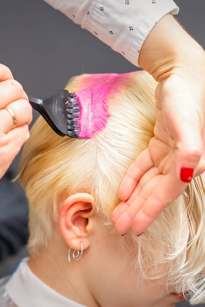 Нанесение розового красителя с расчёской на белые волосы молодой блондинки в парикмахерской - Фото, изображение