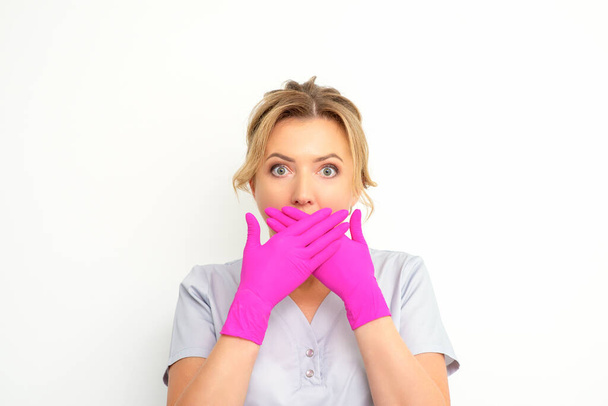 Retrato de una joven doctora o enfermera caucásica sorprendida cubriéndose la boca con sus manos con guantes rosados sobre un fondo blanco - Foto, imagen