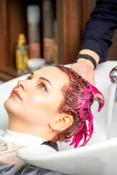 Lavarse el pelo teñido de mujer. Una joven caucásica lavándose el pelo en un salón de belleza. Peluquería profesional lava pintura de color rosa del cabello de un cliente - Foto, imagen