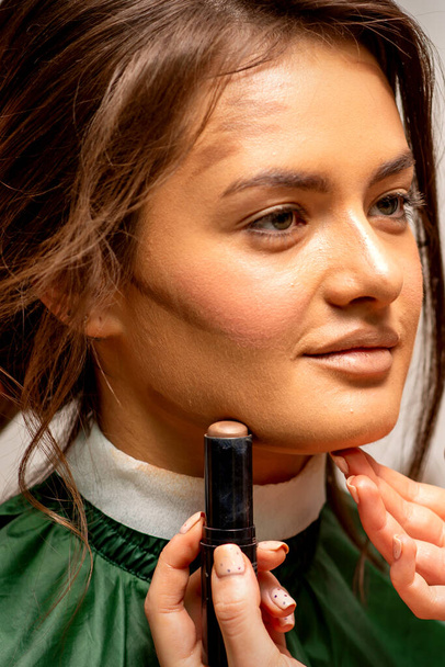 mooi jong brunette vrouw ontvangst make-up met stok concealer op haar gezicht in een schoonheid salon - Foto, afbeelding
