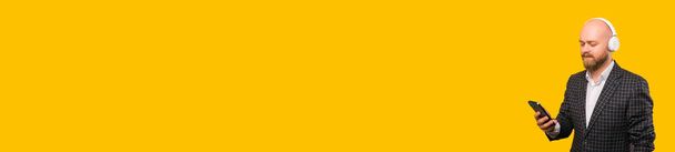 Baner rozmiar zdjęcie brodatego człowieka w luźnym garniturze za pomocą smartfona i noszenia słuchawek na żółtym tle z copyspace . - Zdjęcie, obraz