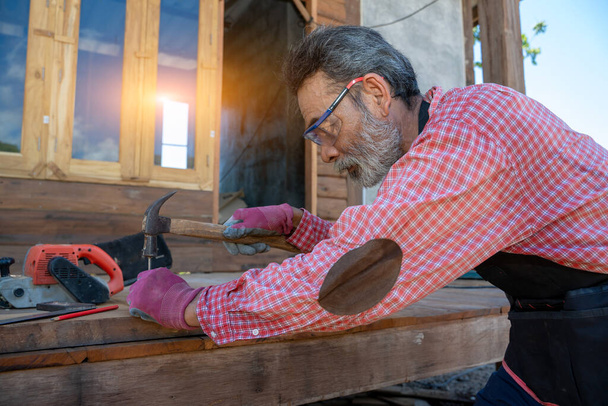 Senior ξυλουργός χρησιμοποιώντας το σφυρί χτύπησε ένα καρφί για συναρμολόγηση ξύλου - Φωτογραφία, εικόνα