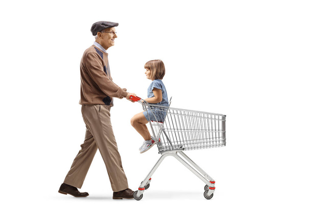 Full length profile shot of an elderly man walking with a child in a shopping cart isolated on white background - Фото, изображение