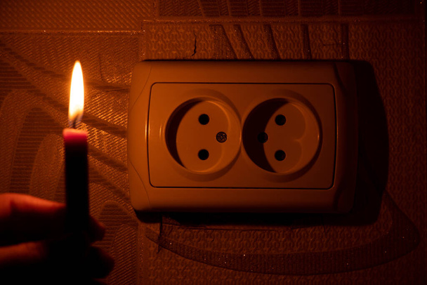 Mano femenina con una vela cerca del enchufe en la pared en el apartamento en la oscuridad, Ucrania, falta de luz debido a la guerra, cortes de energía - Foto, imagen