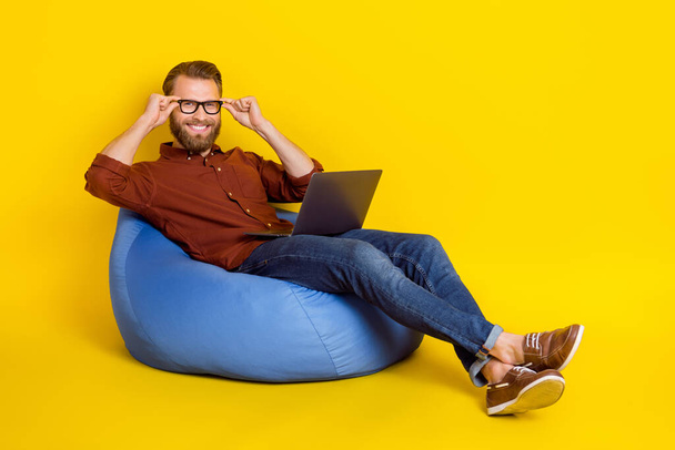 Full body portrait of cheerful person sit cozy bag hands touch eyeglasses use netbook isolated on yellow color background. - 写真・画像