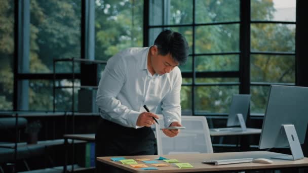 Korean aasialainen työntekijä liikemies seistä pöydällä toimistossa kirjoittaa muistio tarroja hallita projektin tehtäviä. Ammattimainen mies suunnittelija etninen aikuinen japanilainen mies järjestää paperityötä käyttää matkapuhelinta internet - Materiaali, video