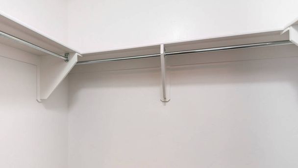 Weiße Regale mit Holzklammern und Metallstangen in einem begehbaren Kleiderschrank. Innenraum eines begehbaren Kleiderschranks mit weißen Wänden und Regalen über dem braunen Teppichboden. - Foto, Bild