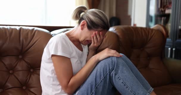 Masentunut nuori nainen peittää kasvonsa käsillään itkien yksin kotona. Järkyttynyt itkevä tyttö istuu sohvalla - Materiaali, video
